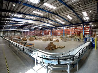 Interior of a warehouse with a conveyor belt, stacks of boxes, and shelves loaded with goods under a high ceiling.
