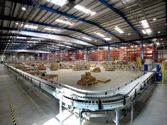 Interior of a warehouse with a conveyor belt, stacks of boxes, and shelves loaded with goods under a high ceiling.