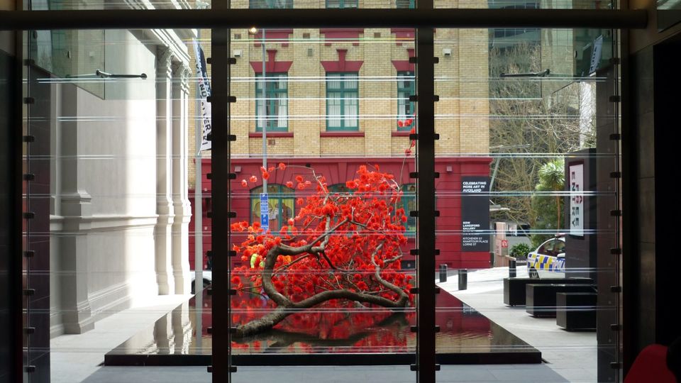 View through large glass windows of a modern lobby featuring a vibrant red art installation outside, contrasted against historic buildings.