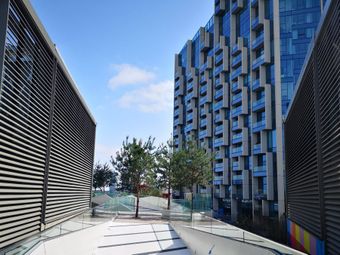 Modern urban walkway in Greenwich flanked by slatted walls, leading to a blue glass building with geometric patterns.