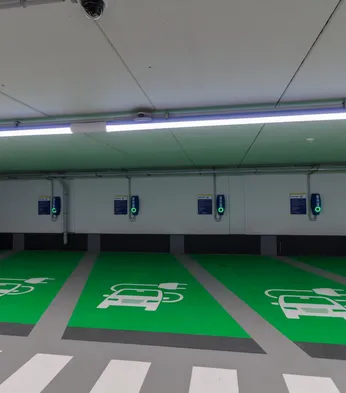 Indoor electric vehicle charging area with green parking spaces, wall-mounted chargers, and car symbols.