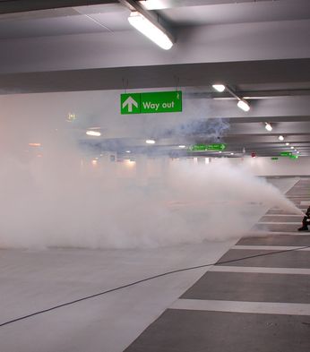 Two people in high-visibility vests operate a fog machine in a parking garage under a "Way out" sign.
