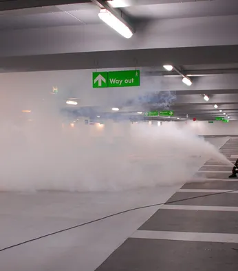 Two people in high-visibility vests operate a fog machine in a parking garage under a "Way out" sign.
