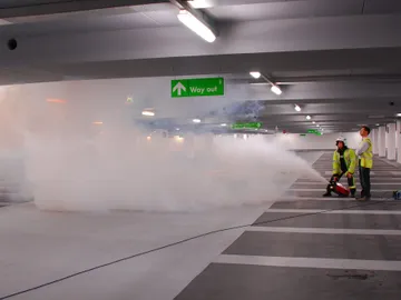 Two people in high-visibility vests operate a fog machine in a parking garage under a "Way out" sign.