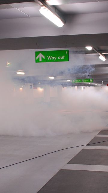 Two people in high-visibility vests operate a fog machine in a parking garage under a "Way out" sign.