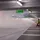 Two people in high-visibility vests operate a fog machine in a parking garage under a "Way out" sign.
