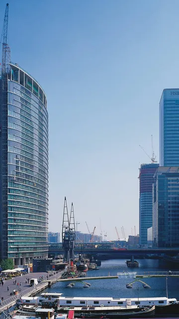 Skyline with modern skyscrapers and a historic building along a river, bustling with people and boats on a clear day.