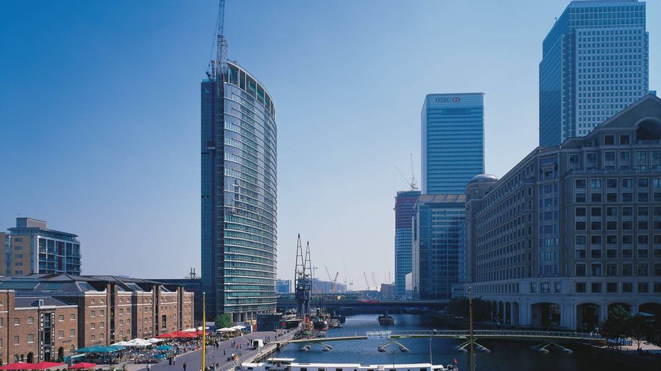 Skyline with modern skyscrapers and a historic building along a river, bustling with people and boats on a clear day.