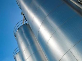 Three large industrial metal silos stand against a clear blue sky, viewed from below, showing their smooth, silver surfaces.