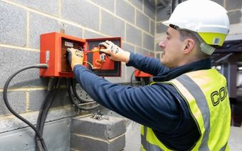 Electrician in a hard hat and high-visibility vest adjusts controls inside a red electrical panel on a brick wall, ensuring system functionality.