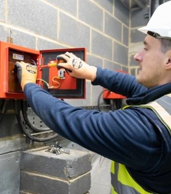 Electrician in a hard hat and high-visibility vest adjusts controls inside a red electrical panel on a brick wall, ensuring system functionality.