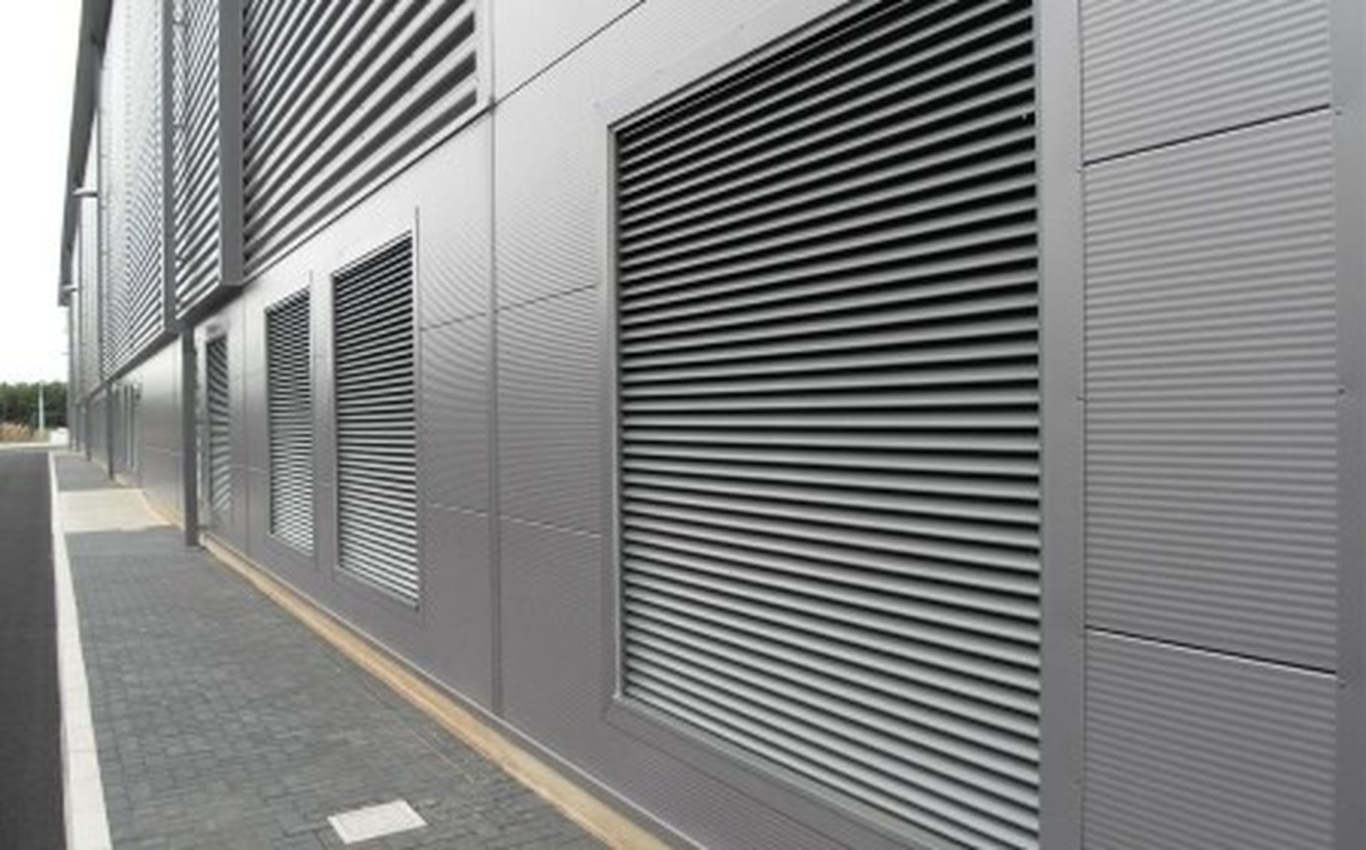 Metal-clad industrial building with large vented panels and parallel-lined corrugated texture, adjacent to a paved pathway.