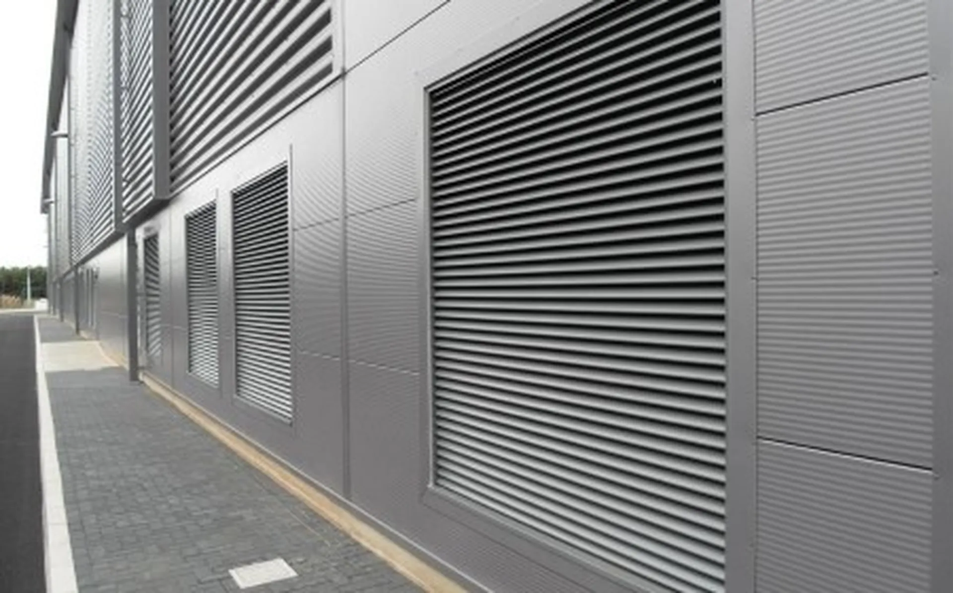 Metal-clad industrial building with large vented panels and parallel-lined corrugated texture, adjacent to a paved pathway.