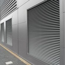 Metal-clad industrial building with large vented panels and parallel-lined corrugated texture, adjacent to a paved pathway.