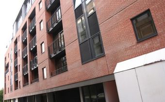 Red brick building facade with multiple windows and small balconies, viewed from a slightly angled perspective.