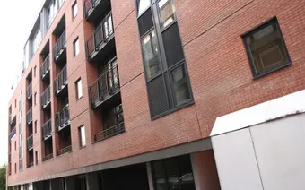 Red brick building facade with multiple windows and small balconies, viewed from a slightly angled perspective.