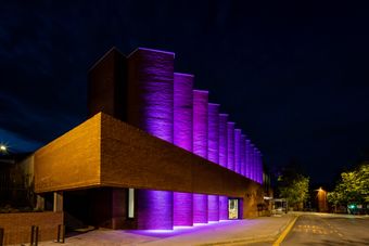 Shakespeare North Playhouse at night illuminated by purple lights, showcasing a zigzag wall pattern; empty road with "bus stop" markings in foreground.