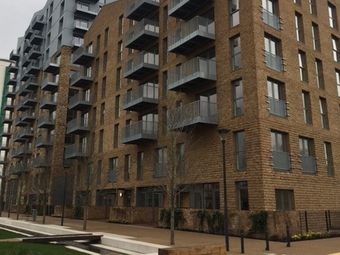 Modern multi-story apartment building with brick facade and numerous balconies, surrounded by a landscaped walkway and bare trees.