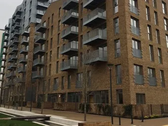Modern multi-story apartment building with brick facade and numerous balconies, surrounded by a landscaped walkway and bare trees.