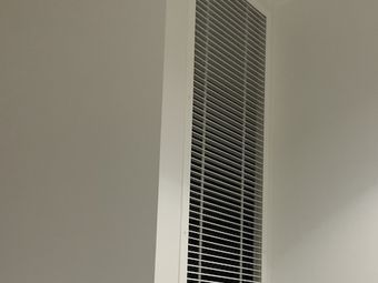 A vertical, rectangular vent with white slats, mounted on a light grey wall.