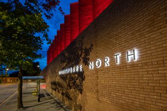A brick building with "Shakespeare North" illuminated on the wall. The roof is lit in red, and a tree casts shadows on the building at dusk.
