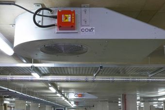Ceiling-mounted ventilation system in a parking garage with metal ducts and a red control box, under fluorescent lighting.