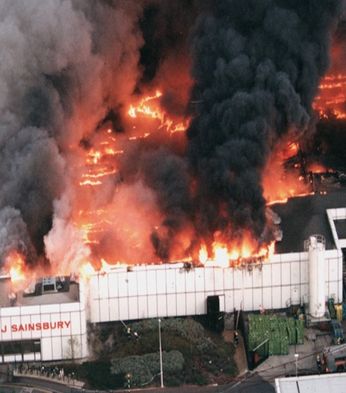 Aerial view of a large supermarket on fire, with flames and thick black smoke billowing from the roof. Fire trucks are positioned nearby.
