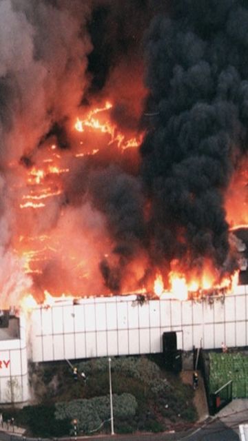 Aerial view of a large supermarket on fire, with flames and thick black smoke billowing from the roof. Fire trucks are positioned nearby.