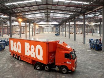 A large orange B&Q truck inside a spacious, empty warehouse with visible columns and ceiling beams. Blue equipment is scattered throughout.