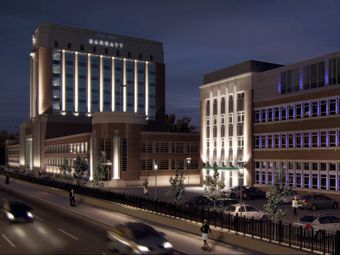A modern, illuminated residential building at night with cars parked out front, next to a busy road with blurred vehicles and pedestrians.