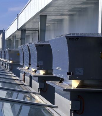 Row of industrial HVAC units on a rooftop, with a cityscape and greenery in the background under a clear blue sky.