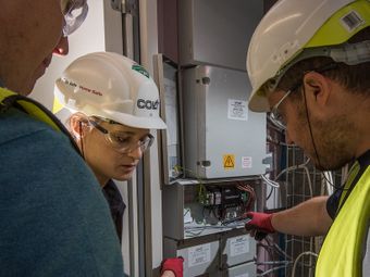Three workers wearing safety gear examine electrical panels in a utility room, with one pointing at the open panel displaying wires and components.