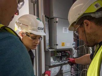 Three workers wearing safety gear examine electrical panels in a utility room, with one pointing at the open panel displaying wires and components.