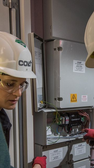 Three workers wearing safety gear examine electrical panels in a utility room, with one pointing at the open panel displaying wires and components.