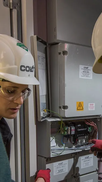 Three workers wearing safety gear examine electrical panels in a utility room, with one pointing at the open panel displaying wires and components.