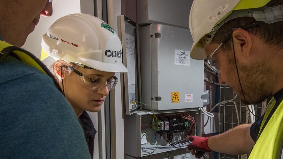 Three workers wearing safety gear examine electrical panels in a utility room, with one pointing at the open panel displaying wires and components.
