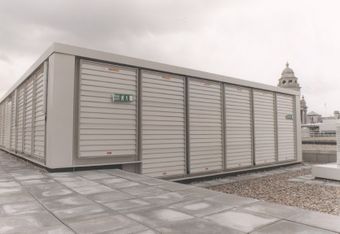 A rooftop ventilation system with louvered panels, with a fire exit sign. Cloudy sky and distant towers are visible in the background.