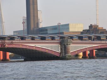 Ornate bridge with red arches and decorative lampposts spanning a river, set against a backdrop of modern and historic buildings.