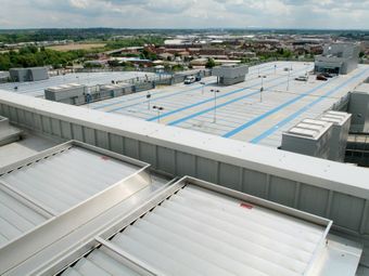 Rooftop view of a large, mostly empty parking structure with blue markings, surrounded by a suburban landscape under a cloudy sky.