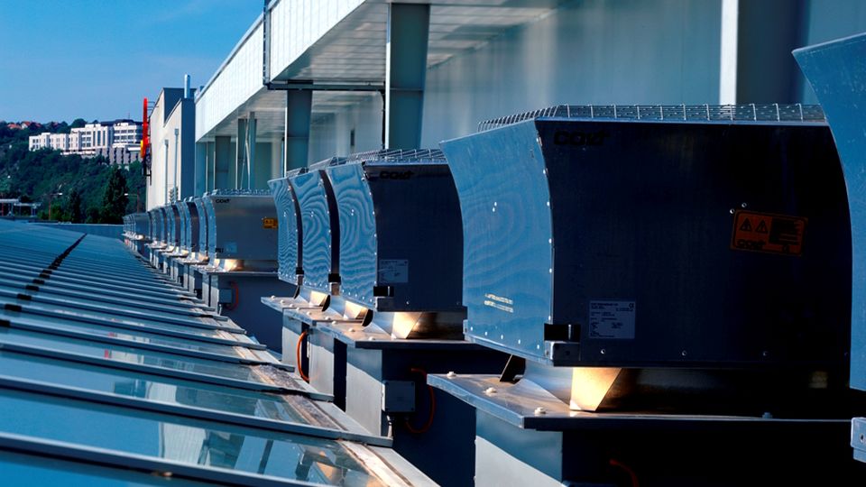 Row of roof mechanical ventilators, with a view of surrounding buildings and greenery in the distance.
