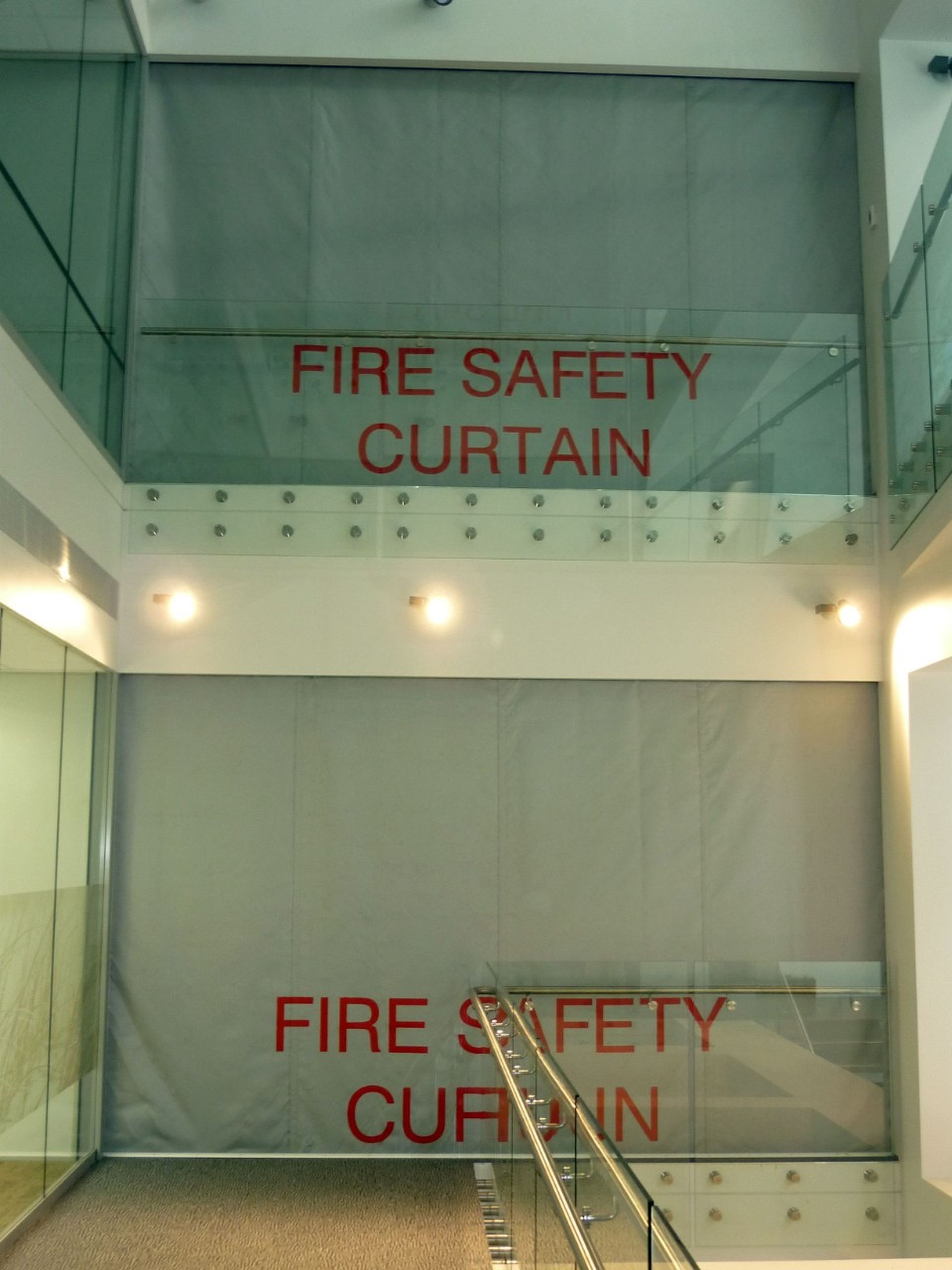 Stairwell with Fire Curtain FM1 deployed on two floors, featuring glass railings and beige walls.