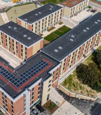 Aerial view of modern, multi-story brick and beige buildings with flat roofs and solar panels, surrounded by green spaces and construction vehicles.