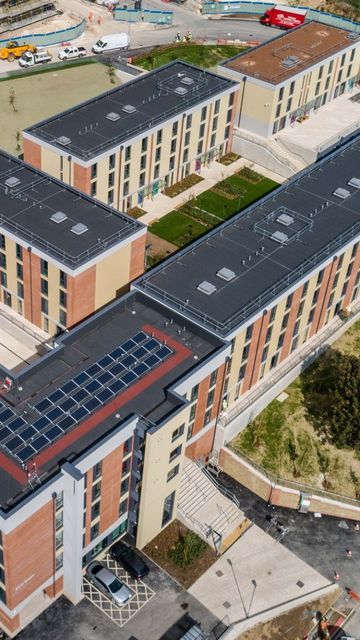 Aerial view of modern, multi-story brick and beige buildings with flat roofs and solar panels, surrounded by green spaces and construction vehicles.