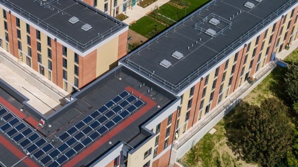 Aerial view of modern, multi-story brick and beige buildings with flat roofs and solar panels, surrounded by green spaces and construction vehicles.