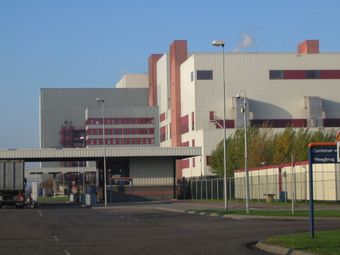 Industrial building with white and red facade, multiple chimneys emitting smoke, surrounded by fencing and road signs, under a clear blue sky.