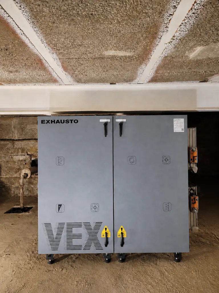 Großes, graues Lüftungsgerät mit den Aufschriften „EXHAUSTO“ und „VEX“, platziert in einem Kellerraum mit strukturierter Decke und unverputzten Wänden.