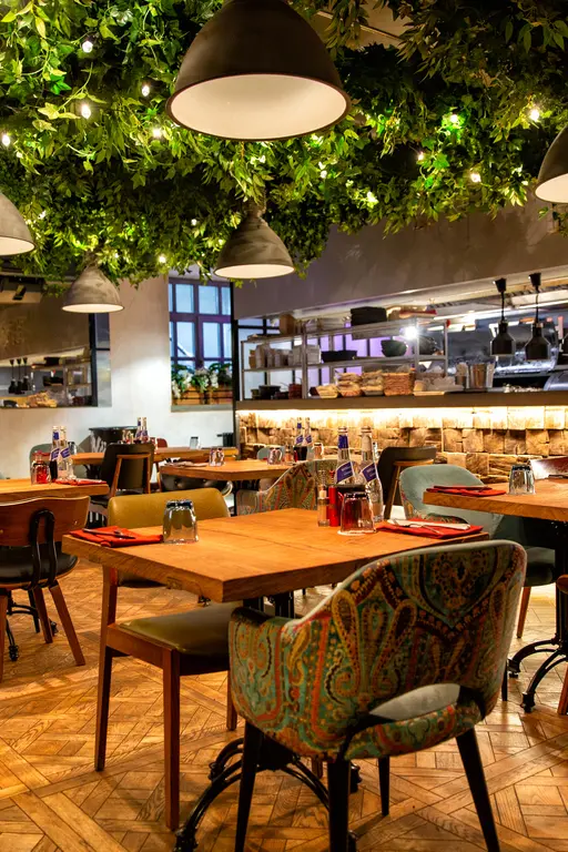 A cozy restaurant interior with wooden tables, colorful chairs, decorative ceiling greenery, pendant lights, and neatly set dining arrangements.