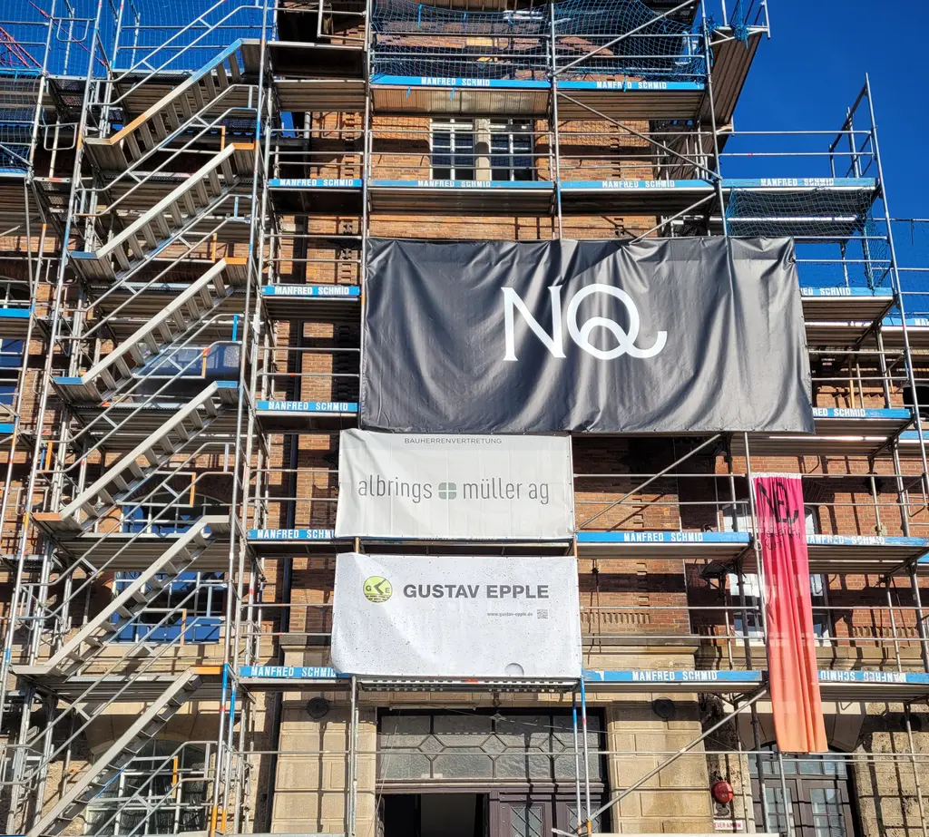 Baustelle eines mehrstöckigen Gebäudes mit vollständiger Fassadengerüstung und Treppentürmen. Im Zentrum ist ein Zugang im Erdgeschoss sichtbar, darüber mehrere Banner mit Firmenlogos, darunter „NQ“, „albrings + muller ag“ und „GUSTAV EPPLE“. Die Fassade besteht aus Backstein und teilweise renovierten bzw. abgedeckten Bereichen. Ein Bauaufzug bzw. Materiallager ist im unteren rechten Bereich erkennbar.