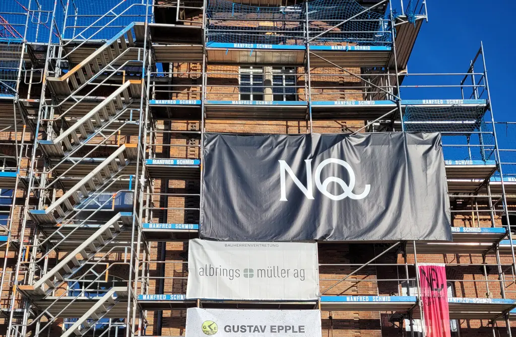 Baustelle eines mehrstöckigen Gebäudes mit vollständiger Fassadengerüstung und Treppentürmen. Im Zentrum ist ein Zugang im Erdgeschoss sichtbar, darüber mehrere Banner mit Firmenlogos, darunter „NQ“, „albrings + muller ag“ und „GUSTAV EPPLE“. Die Fassade besteht aus Backstein und teilweise renovierten bzw. abgedeckten Bereichen. Ein Bauaufzug bzw. Materiallager ist im unteren rechten Bereich erkennbar.