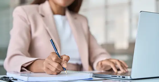 Professional in a pink blazer writing notes with a pen while working on a laptop at an office desk.